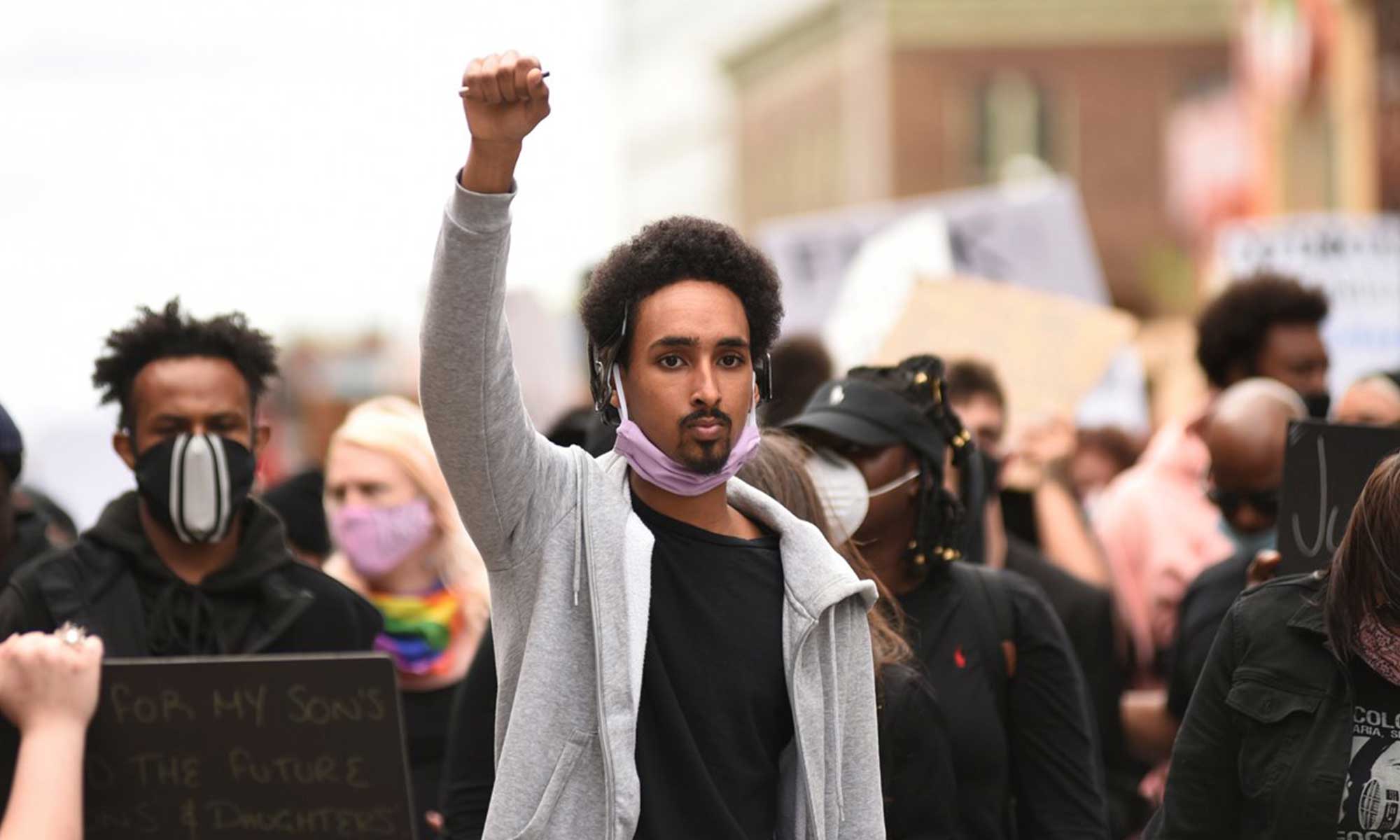 A young black man with an afro and a goatee holds his fist up in the air at a protest.