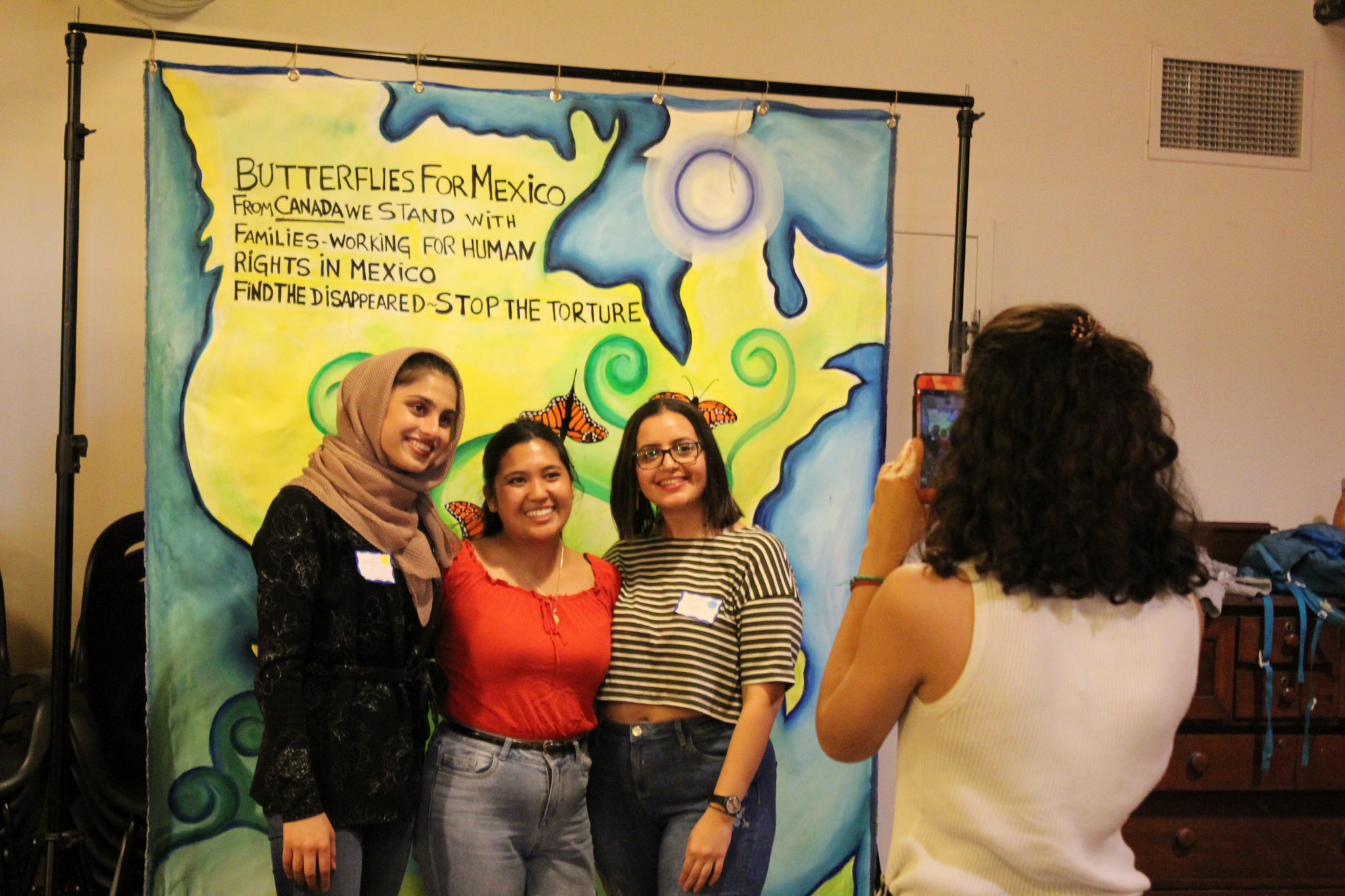 A girl taking a photo on her phone of 3 students standing in front of a painted poster with a map and writing that says "Butterflies for Mexico"