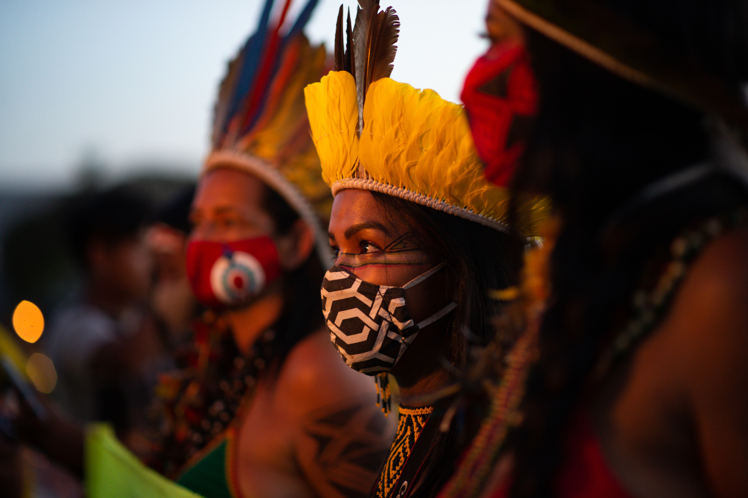 An Indigenous woman wearing a yellow feather headdress and a black and white face stands between two other activists wearing similar attire at a demonstration in Brazil.