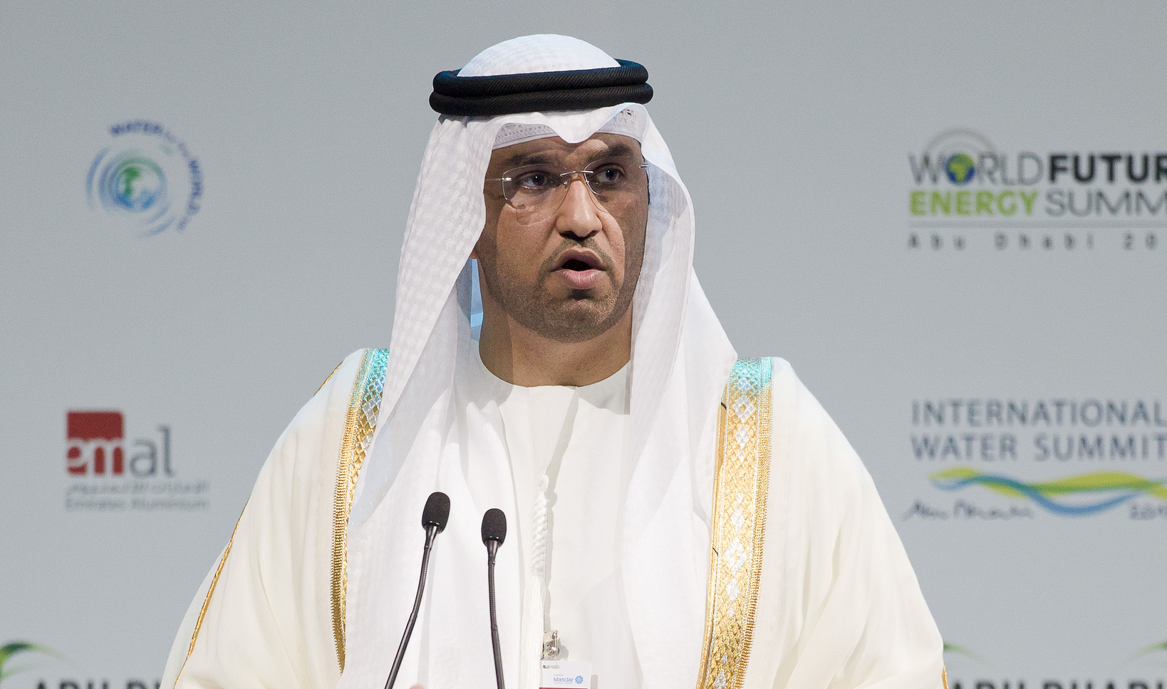 A man wearing a traditional Arab robe called a "thawb" is standing at a lectern giving a speech at a conference.