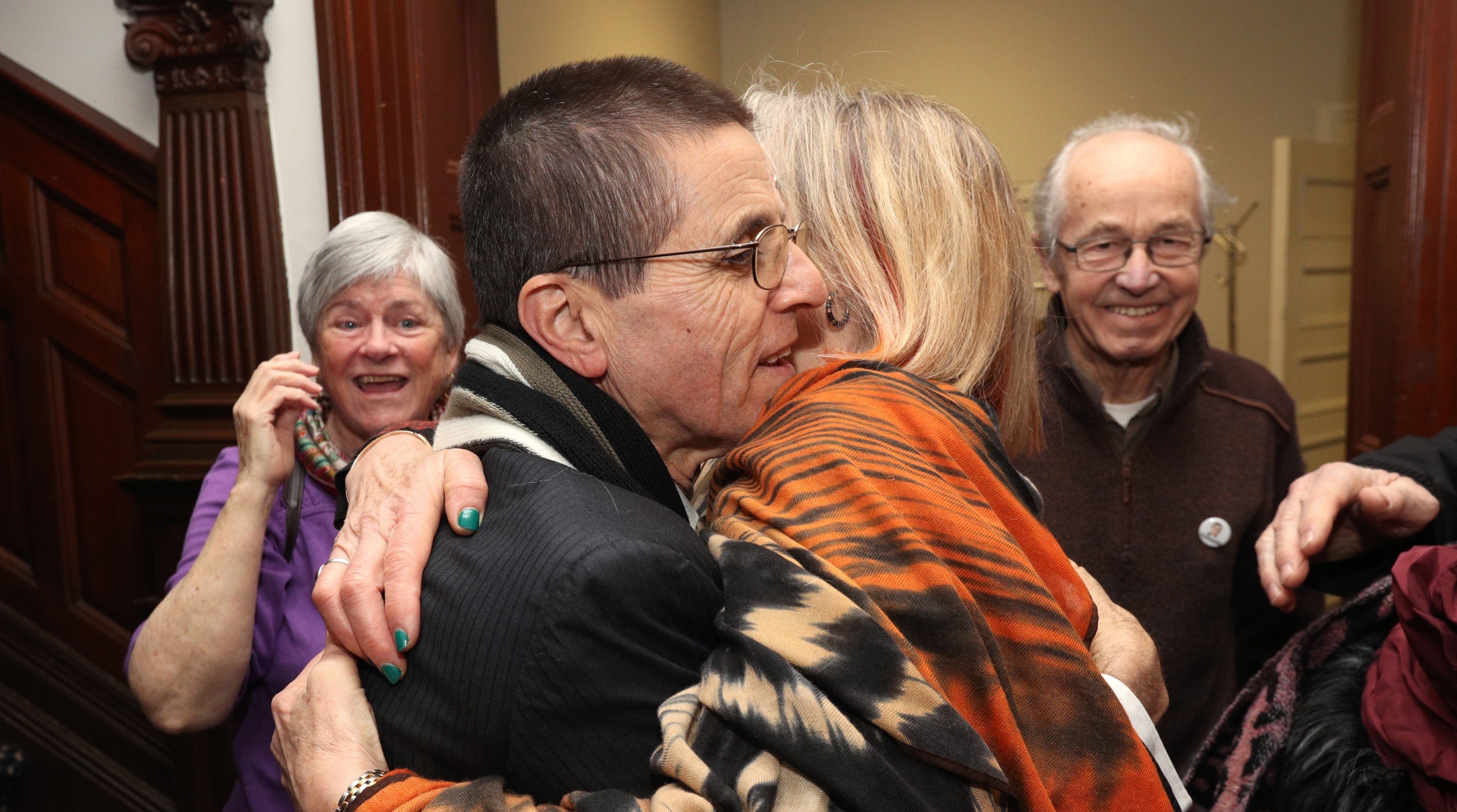 A man in a dark blazer and glasses hugs a woman wearing a flowy black, read and cream-coloured full-length blouse, while two smiling people look on.