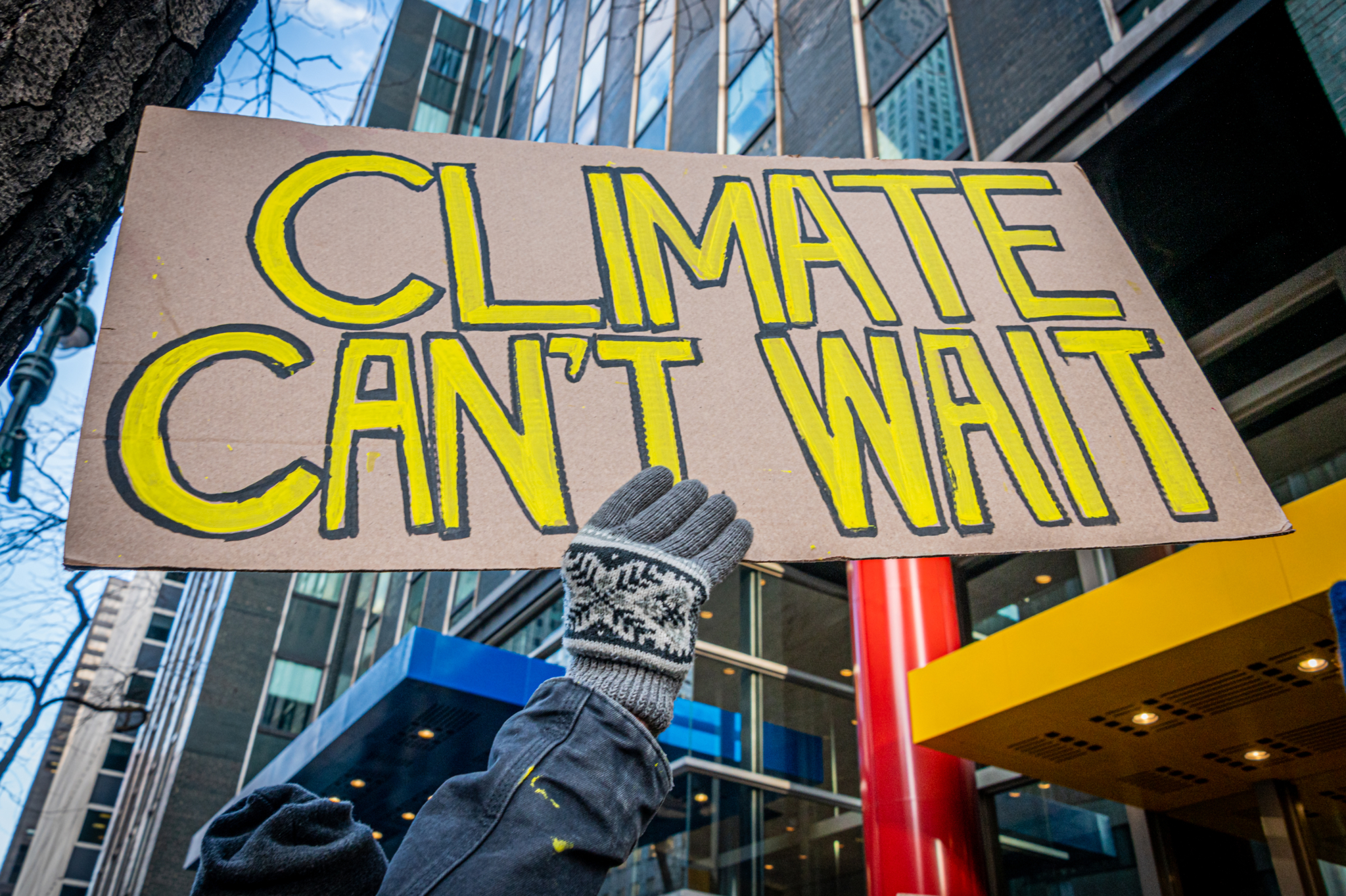 A protester wearing winter gloves hold up a sign that reads "Climate can't wait."