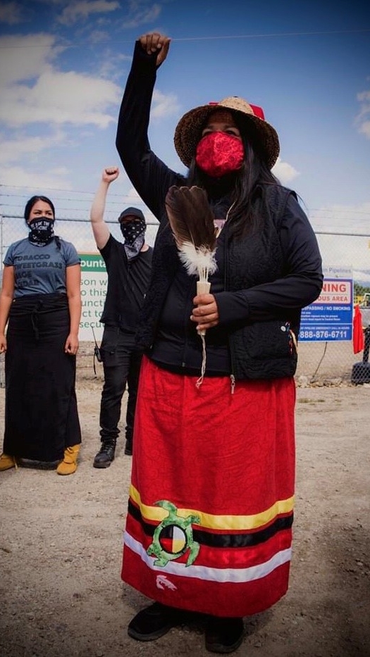 A woman wearing a red face mask, black sweater and a red skirt raises her right fist in the air while holding feathers in her left hand.