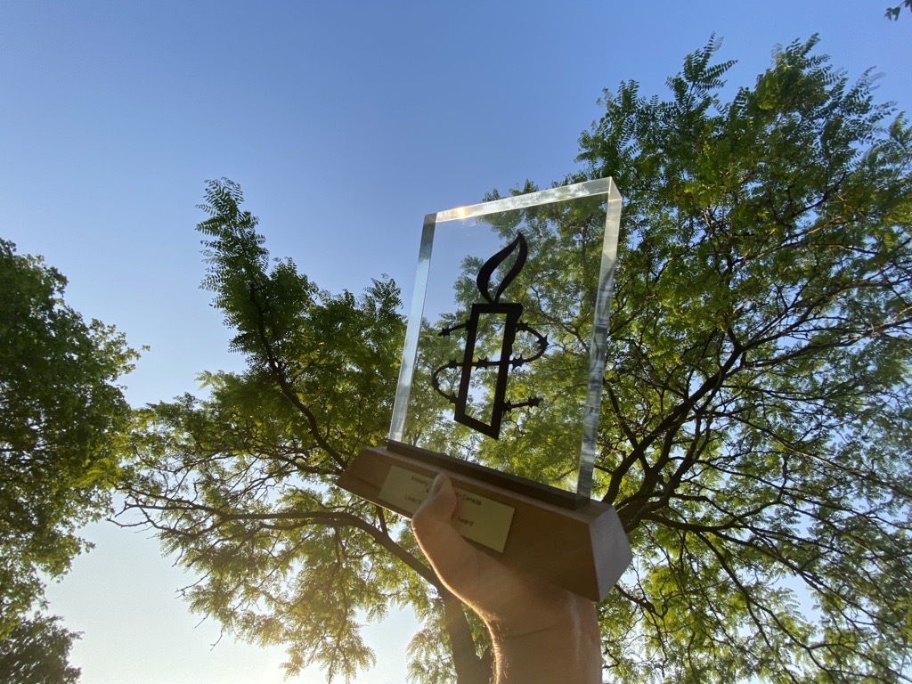 A hand holds an Amnesty Media Awards statue with a wooden base and rectangular, clear glass body with a silhouette of a candle etched into it