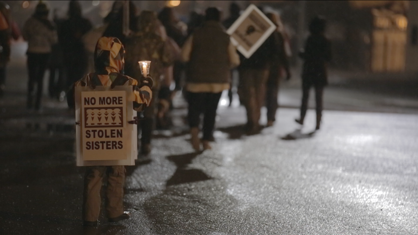 A child holds a sign that reads "No More Stolen Sisters" at a rally on a road at night.
