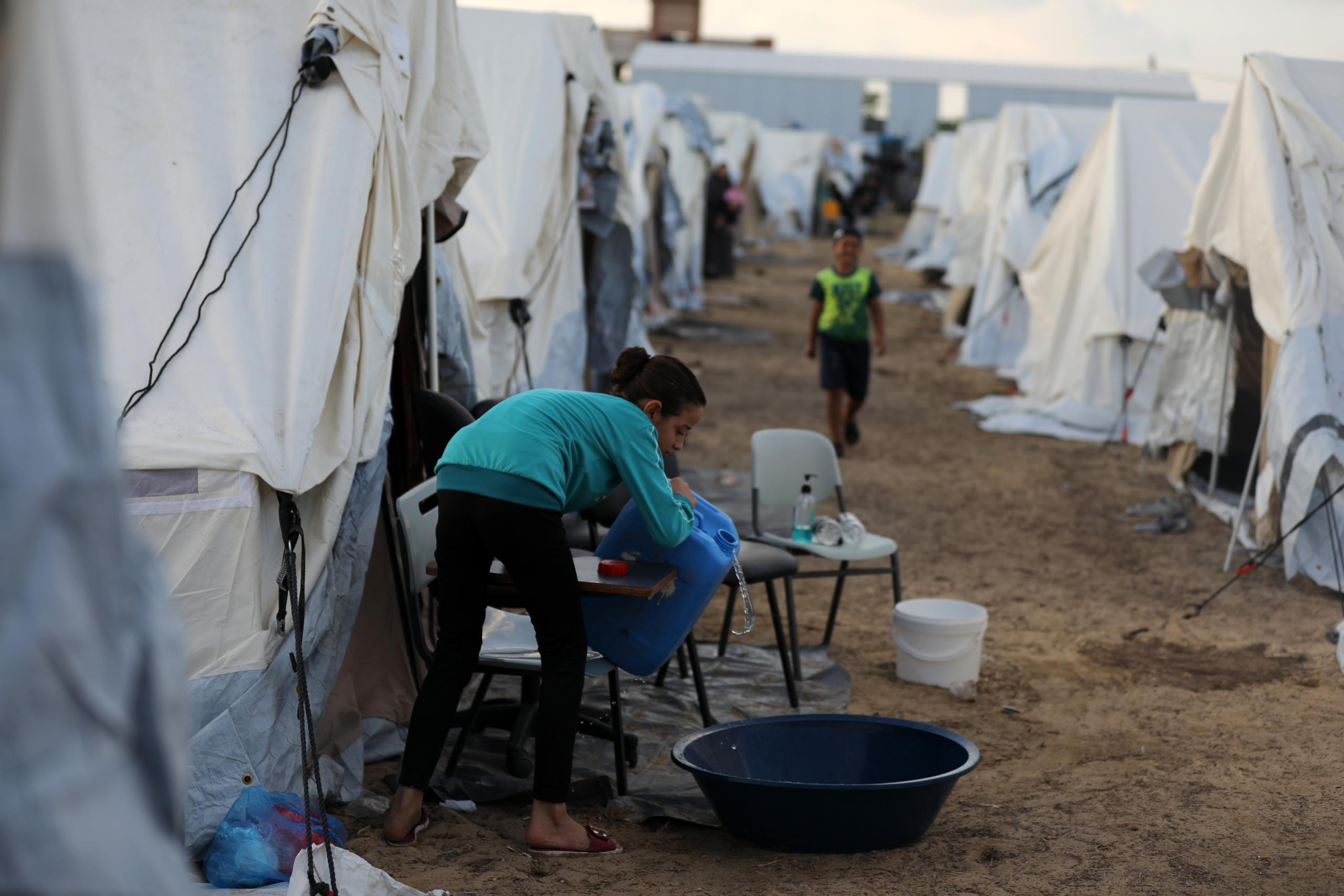 A girl wearing a teal sweater and dark trousers pours water from a jug into a black plastic basin near a row of white tents at what appears to be a refugee camp.