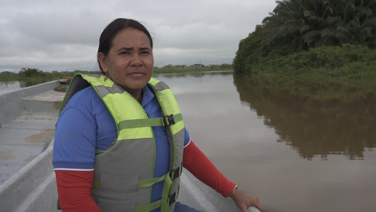A woman sits in a small boat on a river while wearing a blue and read long-sleeve shirt and a neon green and grey life jacket.