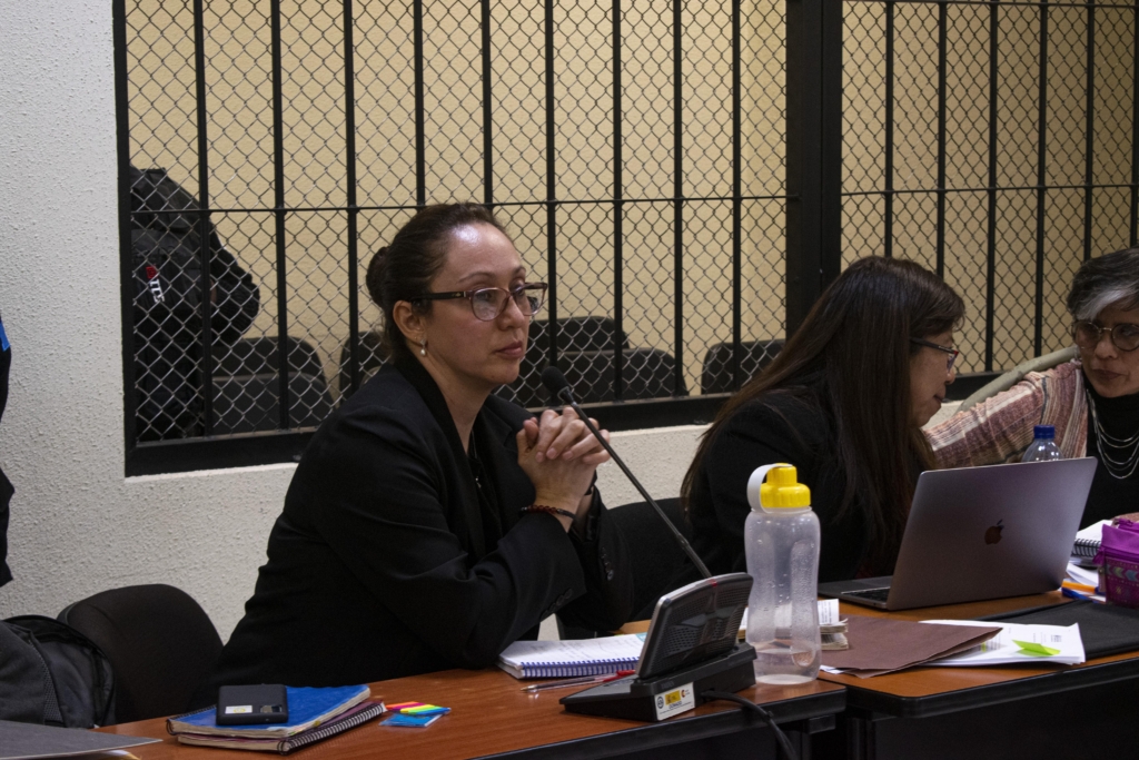 A woman wearing a black blazer and glasses sits with her hands clasped together and her elbows on the desk in front of her.