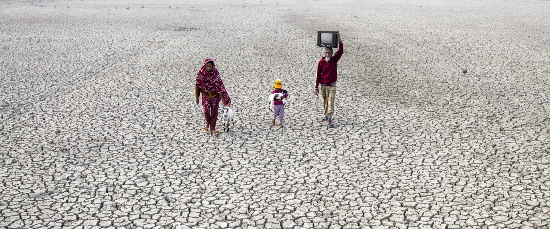 A child carrying a baby goats walks between a man carrying a TV on his head and a woman walking with an adult goat on soil parched by drought.