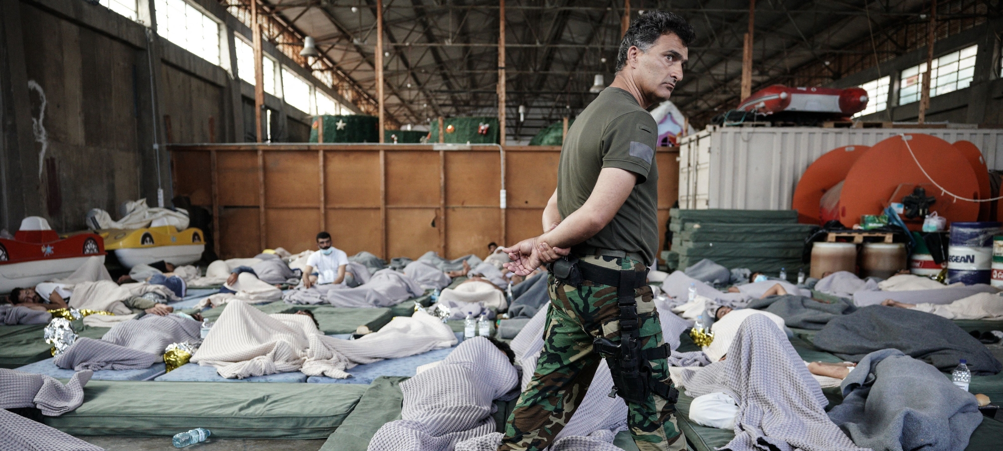 A member of the Coastguard overlooks suvivors as they rest in a warehouse used as a temporary shelter, after a boat carrying dozens of migrants sank in the Ionian Sea, in Kalamata town, Greece, on June 14, 2023. At least 59 people died while more than 100 people have been rescued after the shipwreck. (Photo by Menelaos Myrillas / SOOC / SOOC via AFP) (Photo by MENELAOS MYRILLAS/SOOC/AFP via Getty Images)