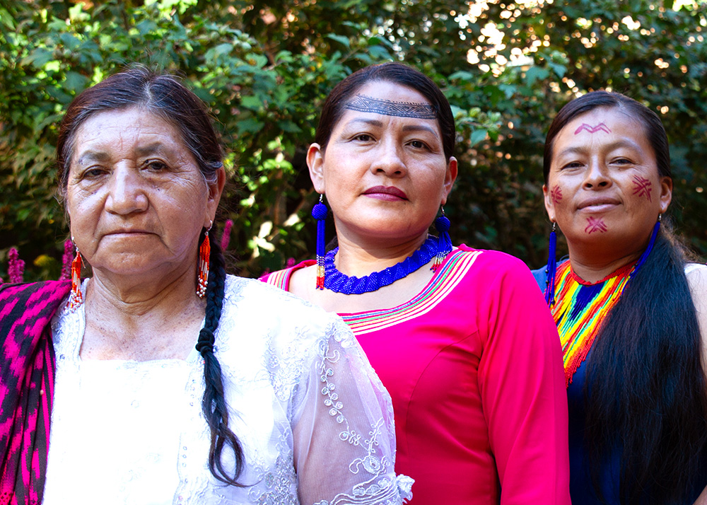 Three women Indigenous and environmental rights defenders pose outdoors in front of a garden