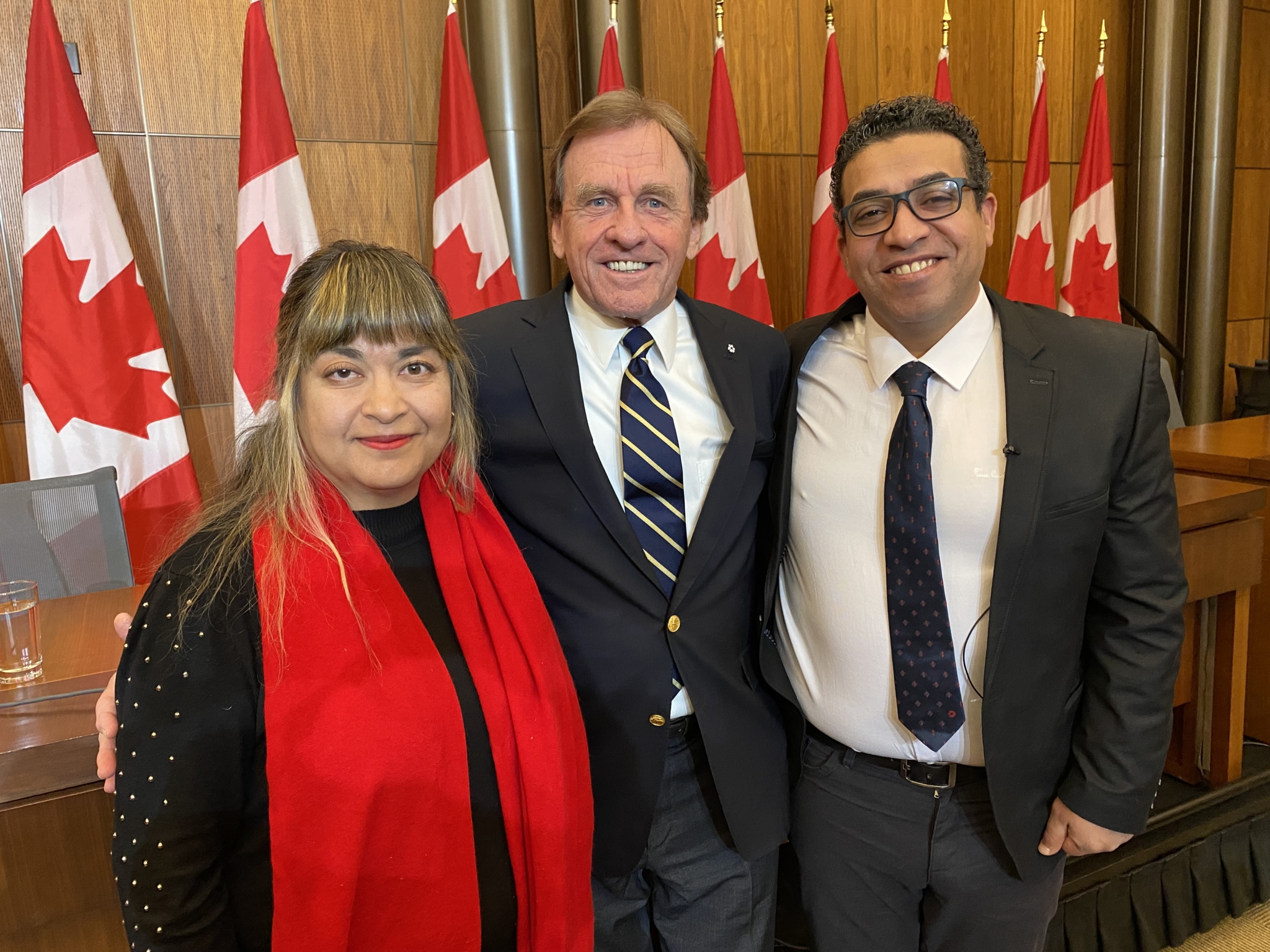 Three people, a woman in a black sweater and red scarf, and two men in dark suit jackets with dark ties, pose in front of a row of nine Canadian flags.