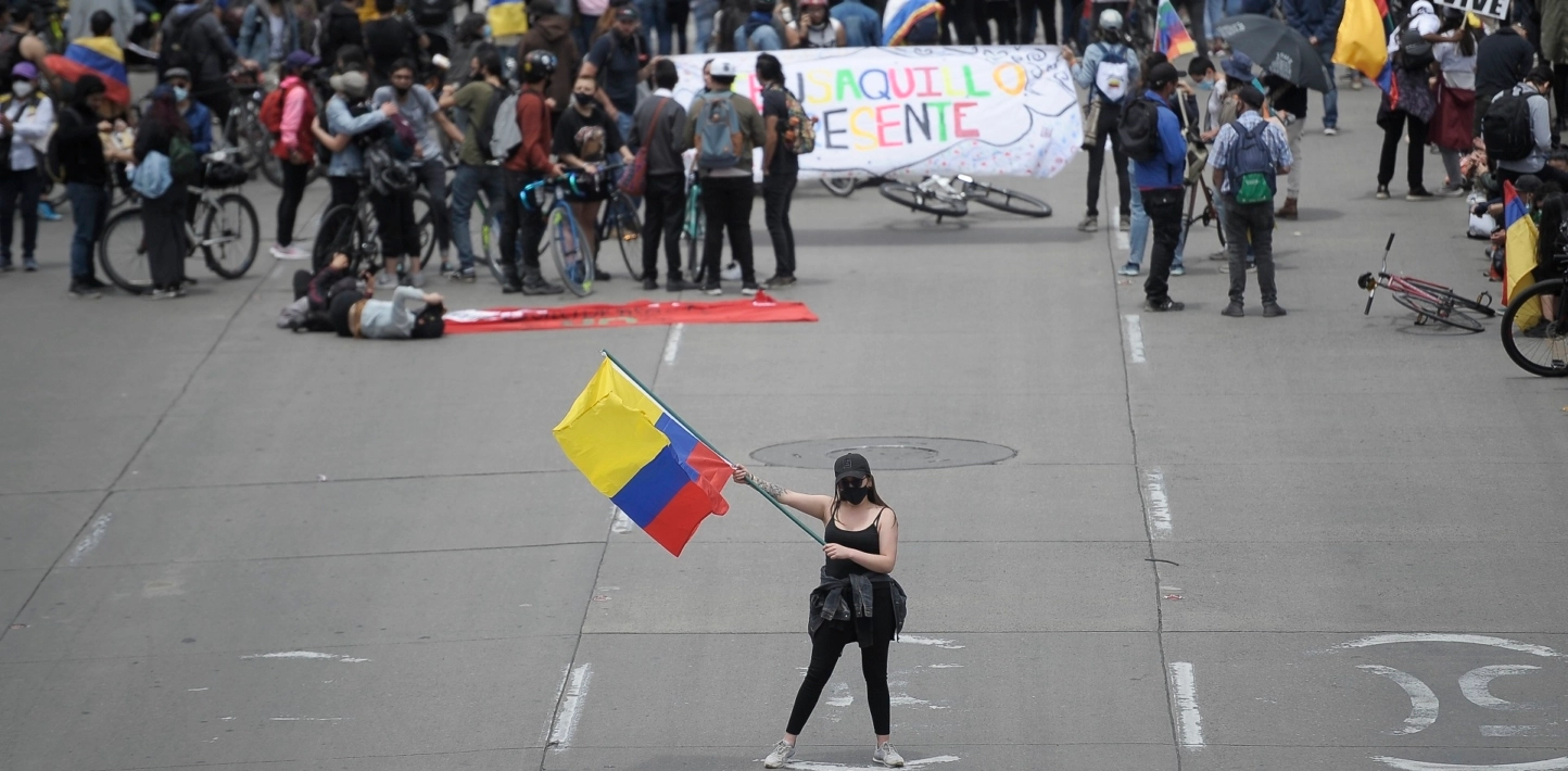 A woman wearing a black face mask and a black tank top and tights waves a Colombian flag in the middle of a large street during a demonstration, while other protester march behind her.