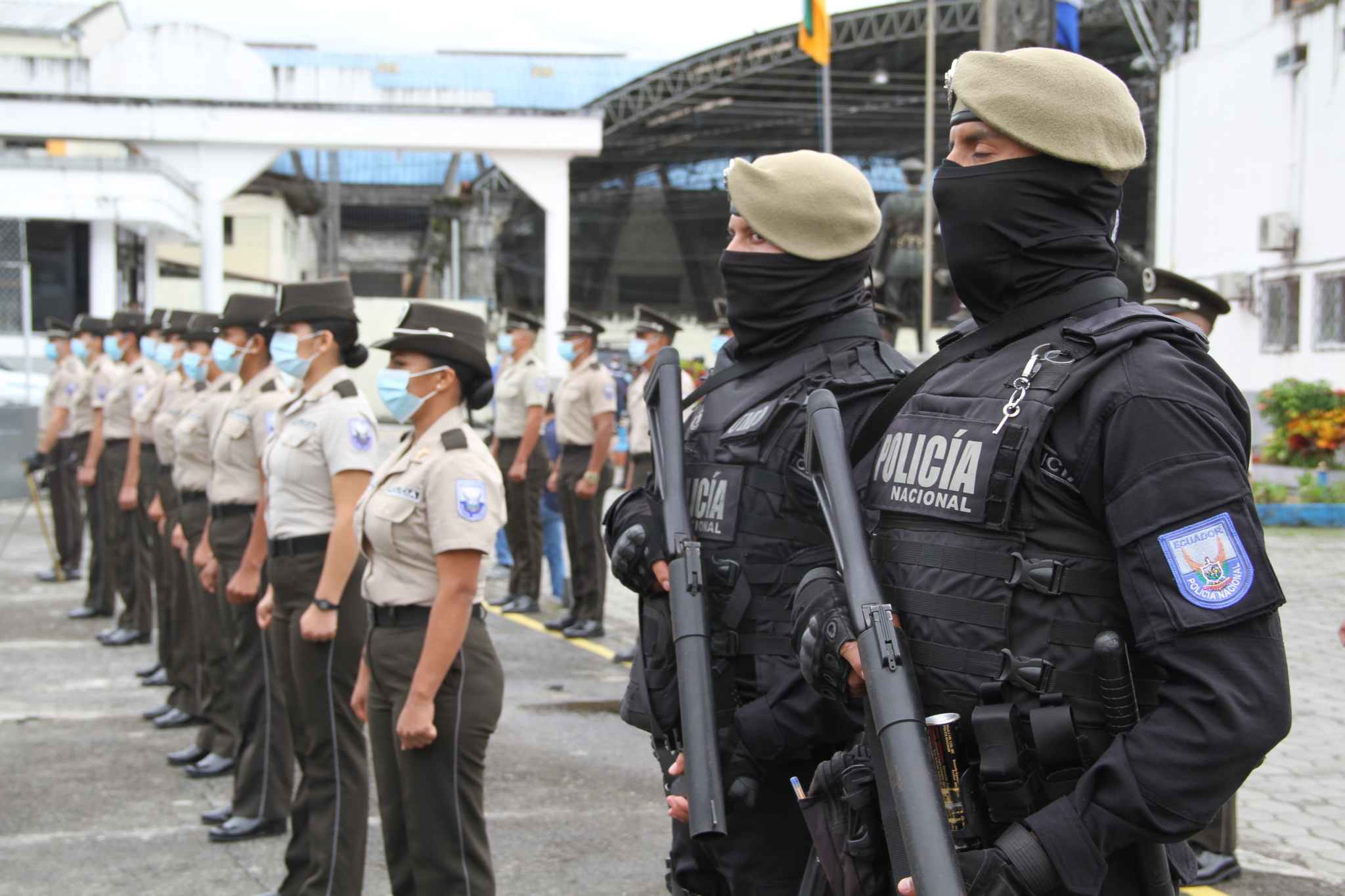 Two police officers carrying heavy guns and wearing and black bulletproof vests and body armour stand beside of row of women police officers wearing dark green slacks and short-sleeve khaki shirts.