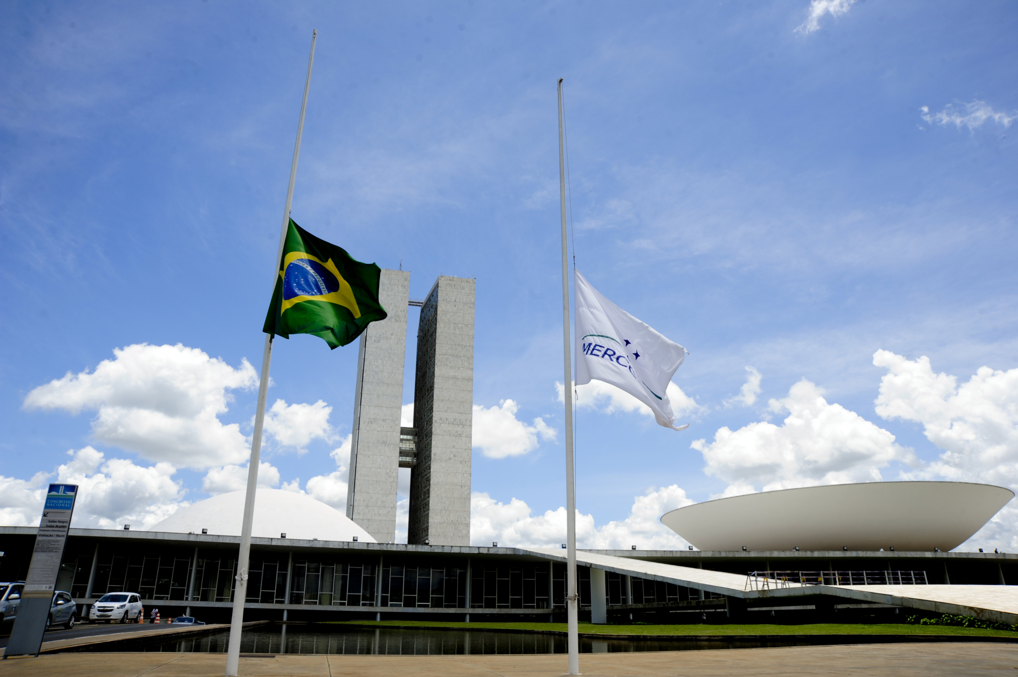 A green, blue and yellow Brazilian flag and a white Mercosur flag stand at half mast in front of a concrete building on a sunny day with partially cloudy skies.