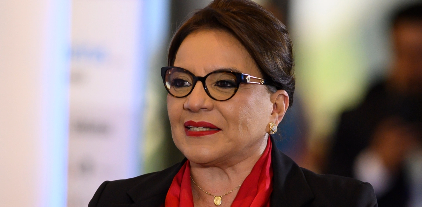 A middle-aged women with black-rimmed cat-eye glasses, brown hair that is parted in the centre, a black blazer, and a red, collared blouse smiles.