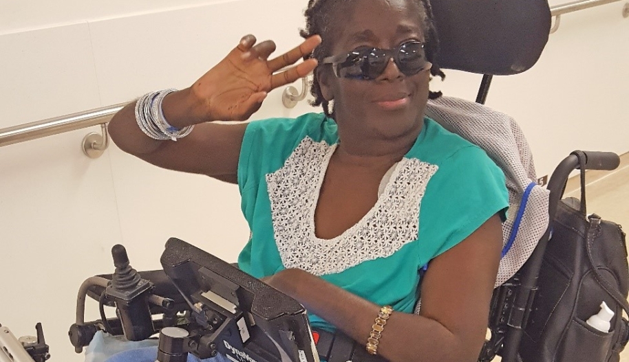 A women in a black electric wheelchair wearing sunglasses, a teal t-shirt with a lacy white collar and lavender-coloured trousers raises her right hand to her forehead as a salute.