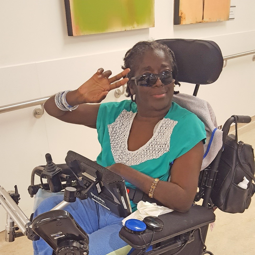 A women in a black electric wheelchair wearing sunglasses, a teal t-shirt with a lacy white collar and lavender-coloured trousers raises her right hand to her forehead as a salute.