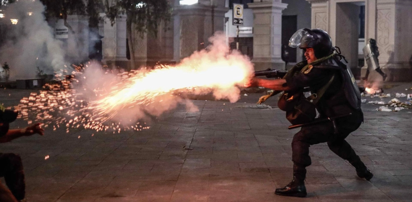 A police officer wearing a black helmet and black body armour fires a flare while crouching in a forward lunge position.