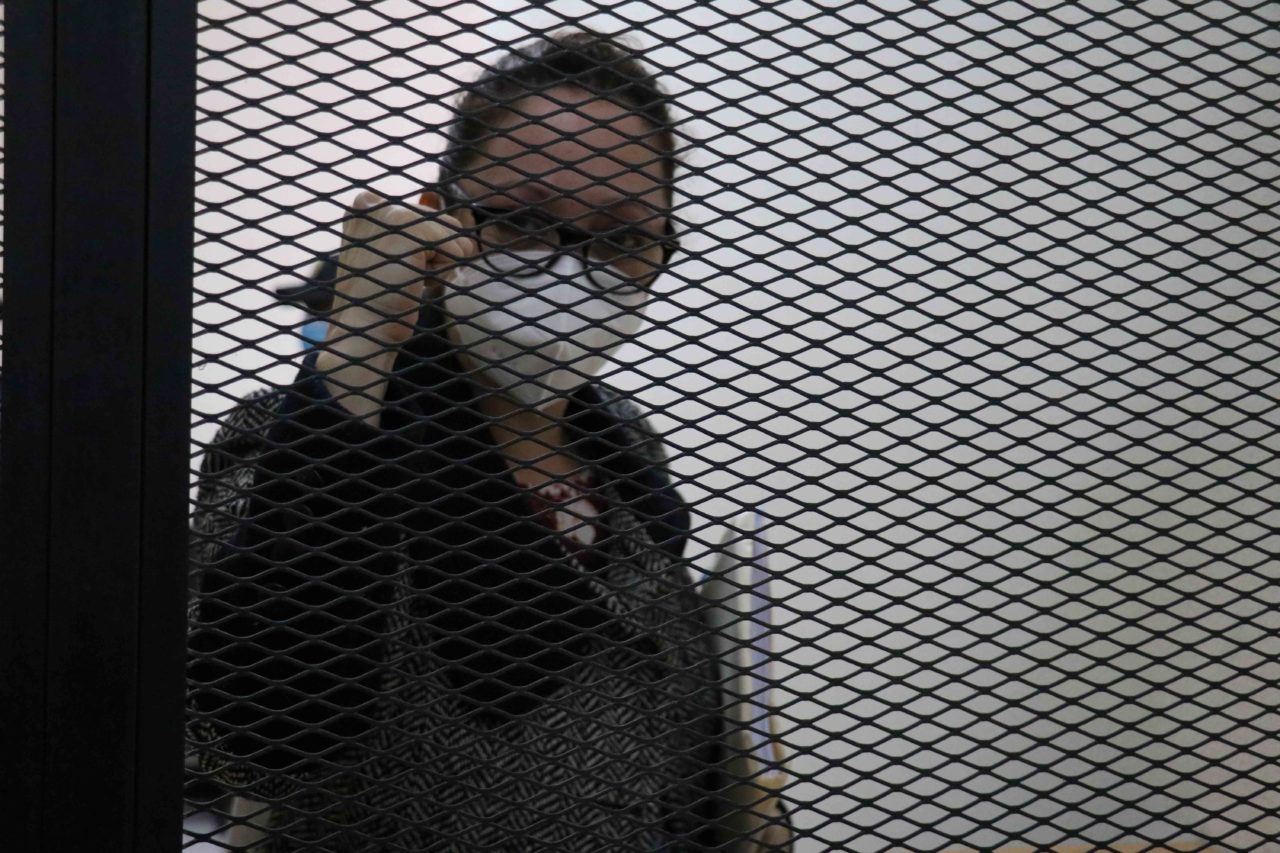 A woman in a dark suit (Virginia Laparra) presses her fist to a window that is re-enforced by a metal grate.