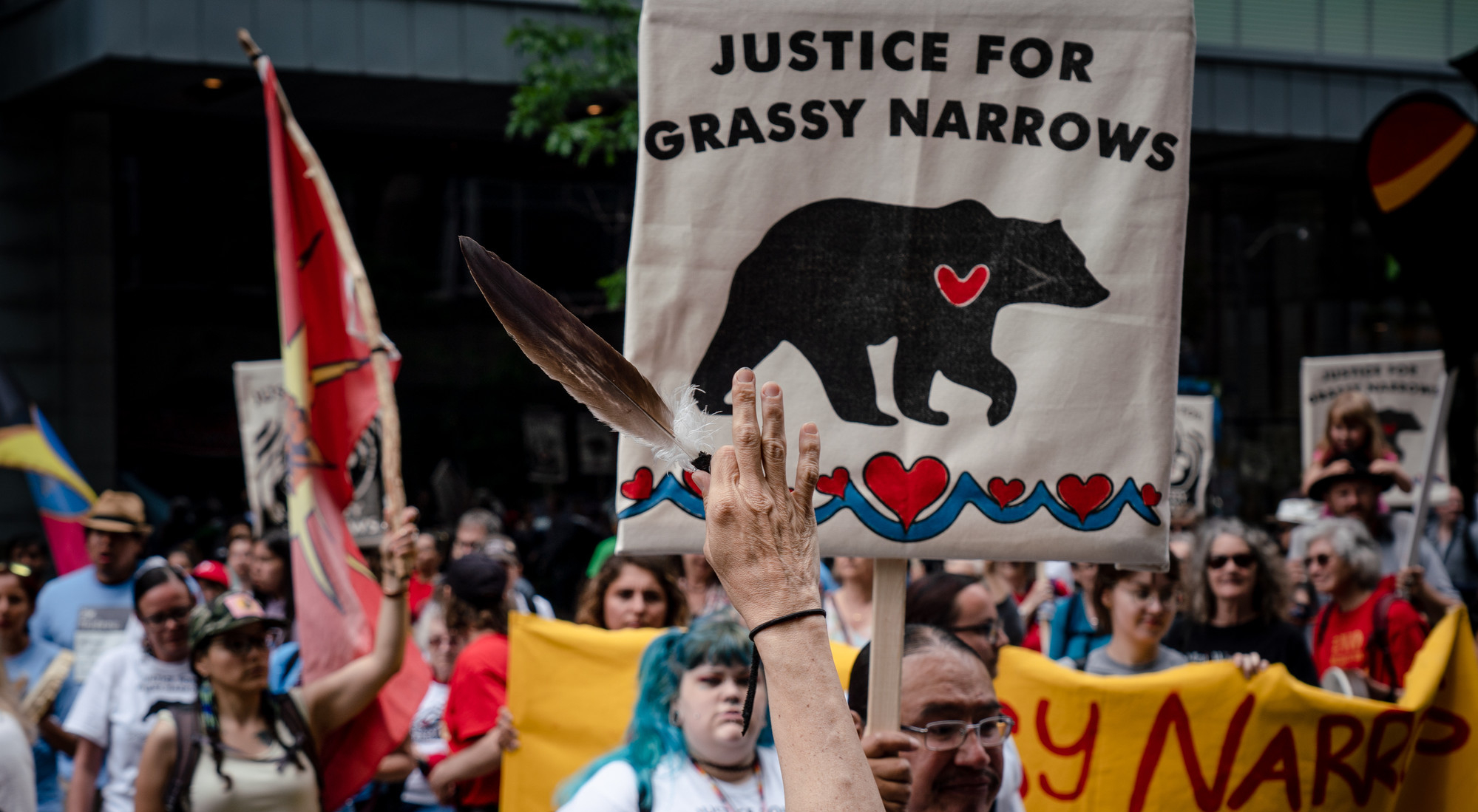 A hand holds up a feather at an Indigenous rights protest in front of a man holding a sign that says "Justice for Grassy Narrows."