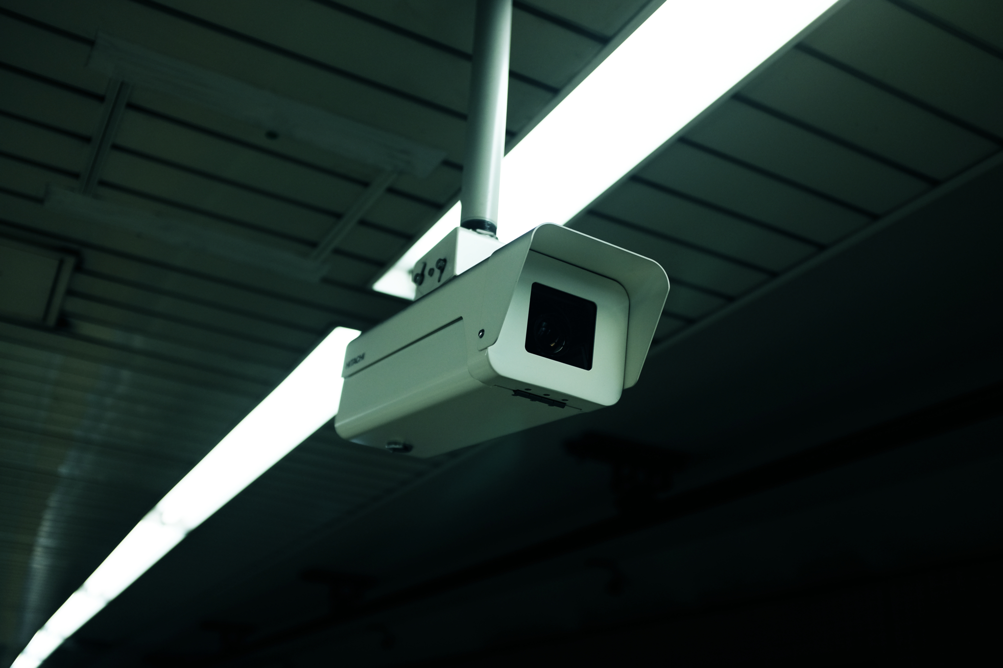 White surveillance camera monitoring a dark space with lined ceiling and overhead lights