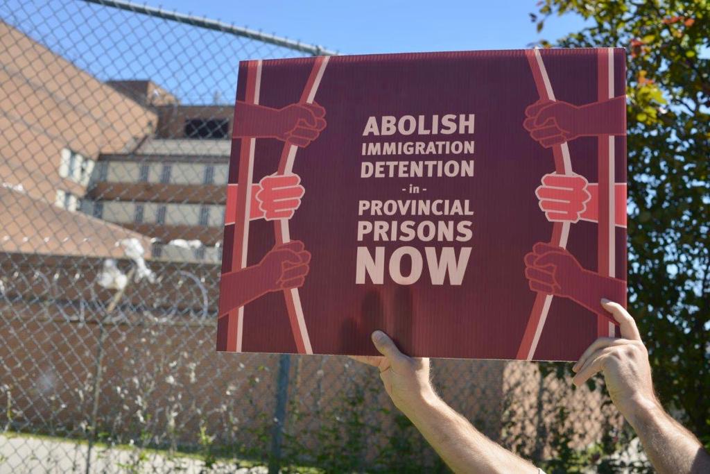 Two hands hold up a sign that reads, "Abolish Immigration Detention in Provincial Jails Now."