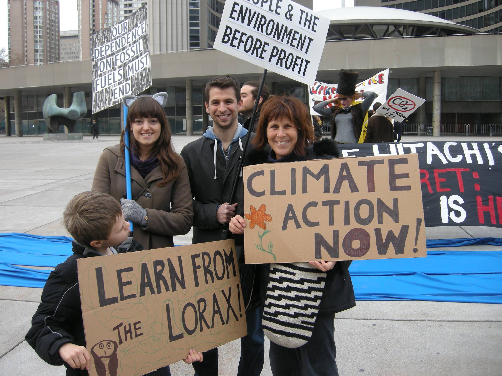 Smiling protesters carry signs that say "Climate Action Now", "Learn From The Lorax" "Our Dependence on Fossil Fuels Must End", "People & the Environment Before Profit"