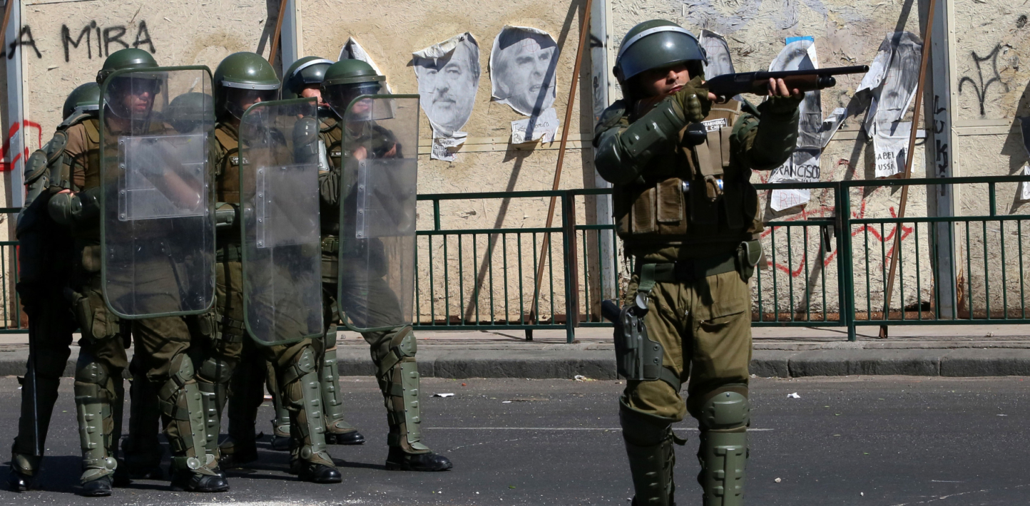 A riot police officer, standing in front of six other riot police officers standing behind shields, fires rubber bullets at demonstrators.