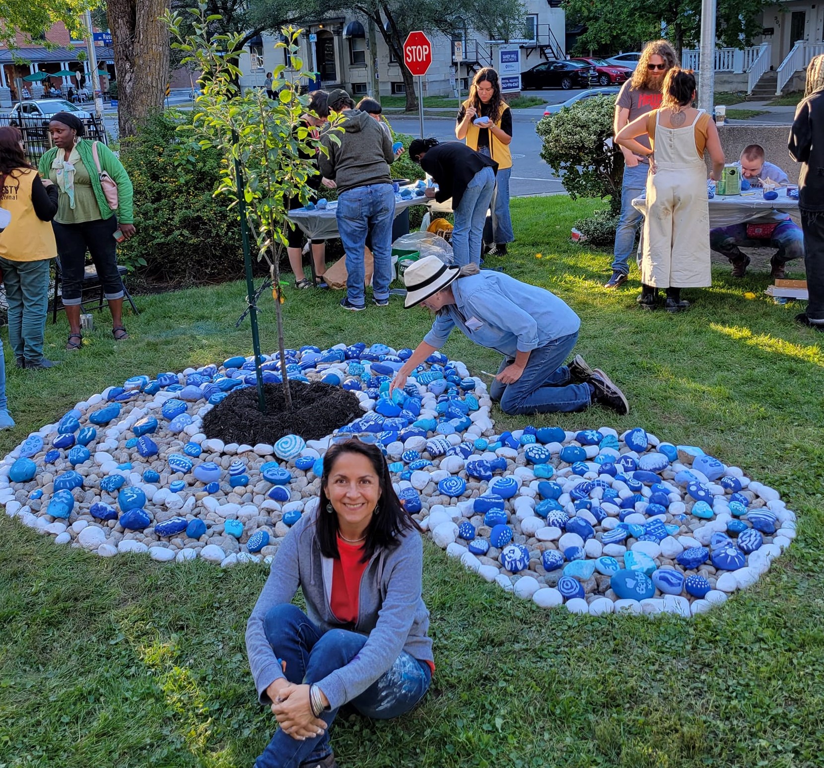 People paint blue and white stones stones to add to a swirling "river" of stones around a tree.
