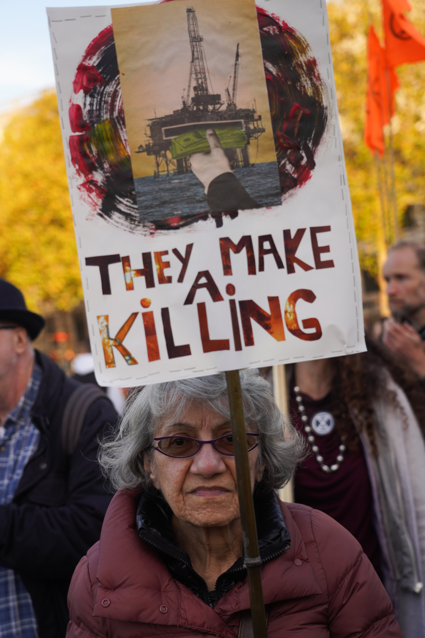 An older woman holds a sign with an image of an oil rig, a stack of money, and the words: "THEY MAKE A KILLING"