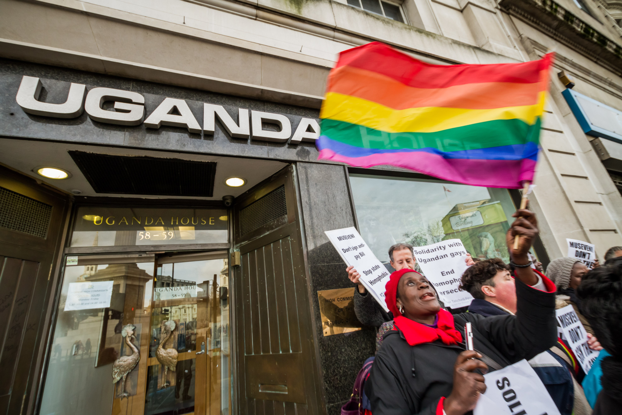 A woman with black jacket, a red had and a red scarf waves a rainbow flag at a protest in in front of a building labelled "Uganda House" on its front door.