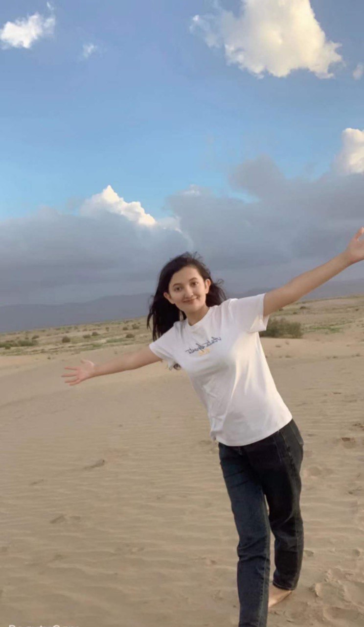 Kamile Wayit pictured on the beach wearing a white t-shirt and black pants with arms wide open.