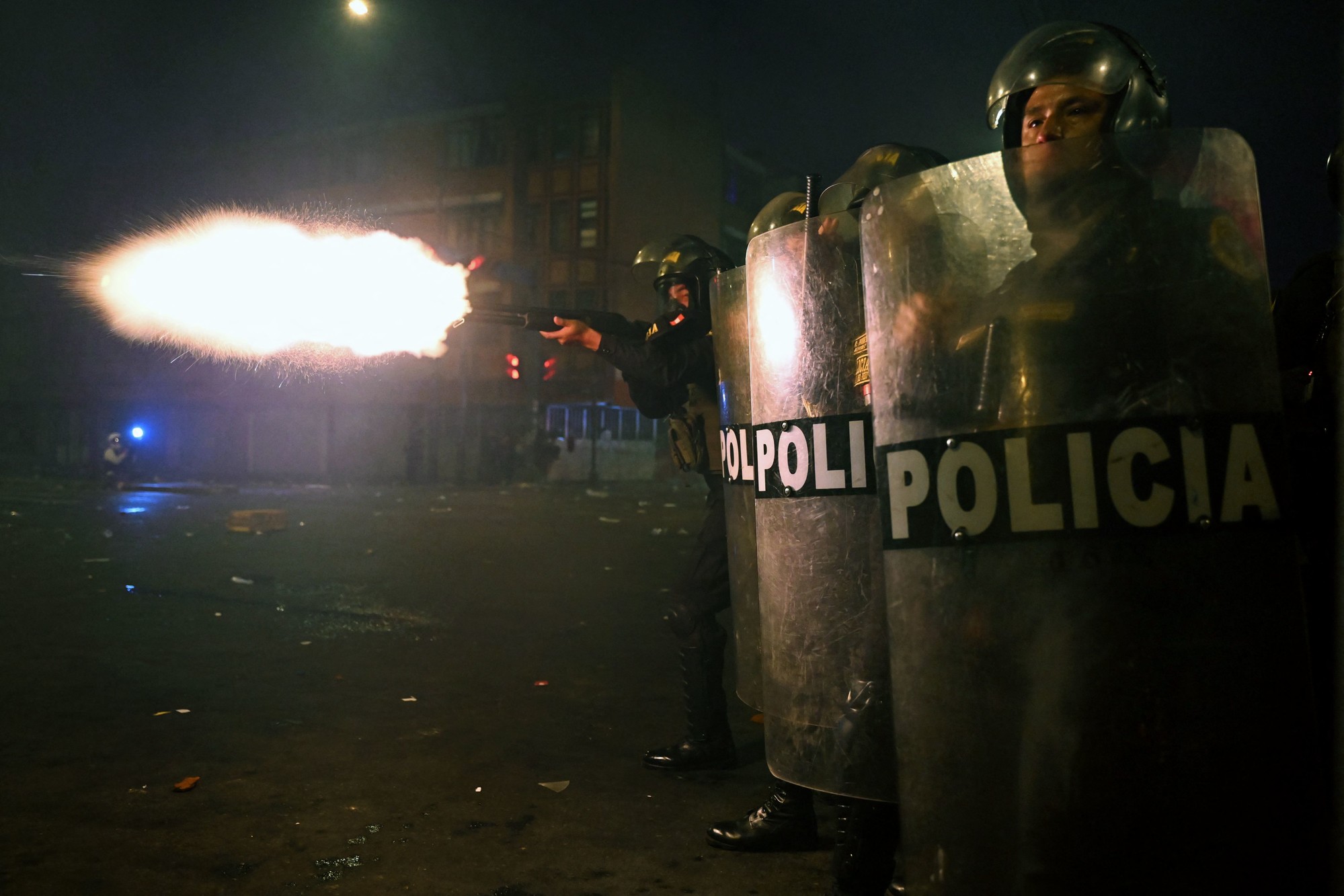 Two police officers in riot gear hold shields while another police officer behind them fires a gun.