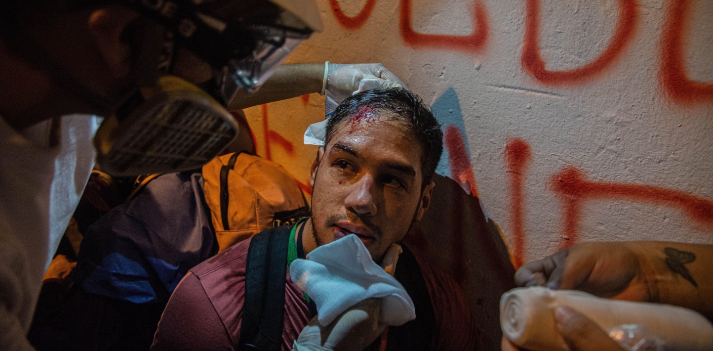 A young man with a wound on his head is leaning against a wall while a medic treats his injury.
