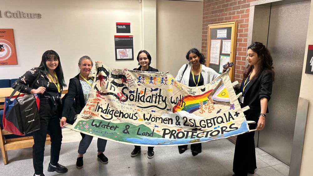 Five women hold up a banner that reads "In Solidarity with Indigenous Women and 2SLGBTQIA+ Water and Land Protectors."