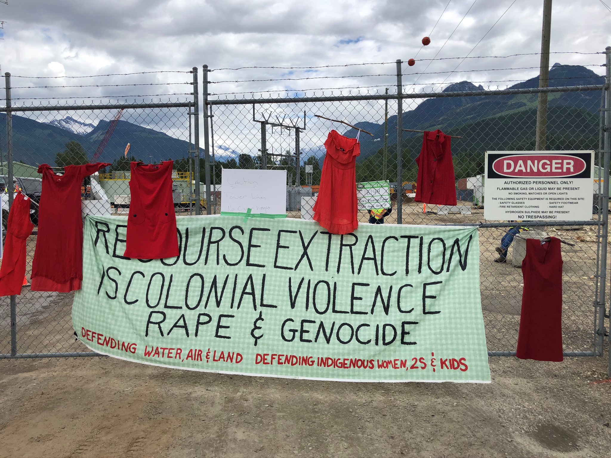 A protest banner tied to a barbed-wire fence reads "Resource extraction is colonial violence, rape and genocide."