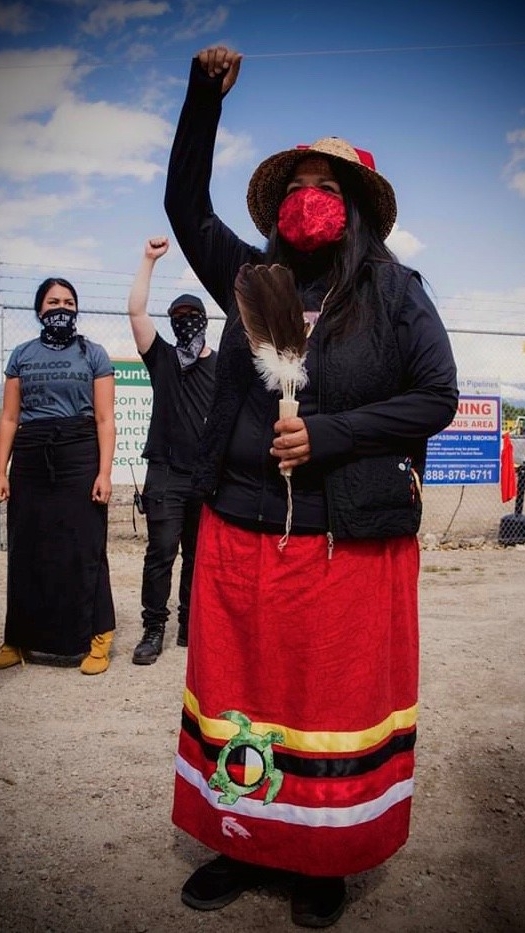 A woman wearing a red face mask, black sweater and a red skirt raises her right fist in the air while holding feathers in her left hand. 