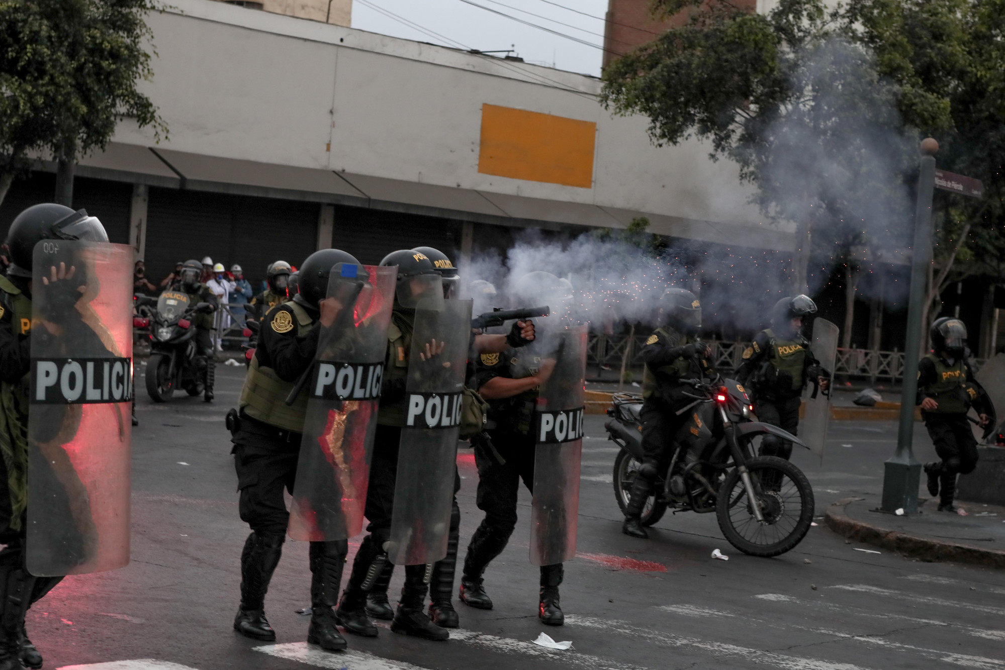 Four police in riot gear hold full-body-length shields and fire what looks like tear gas towards protesters