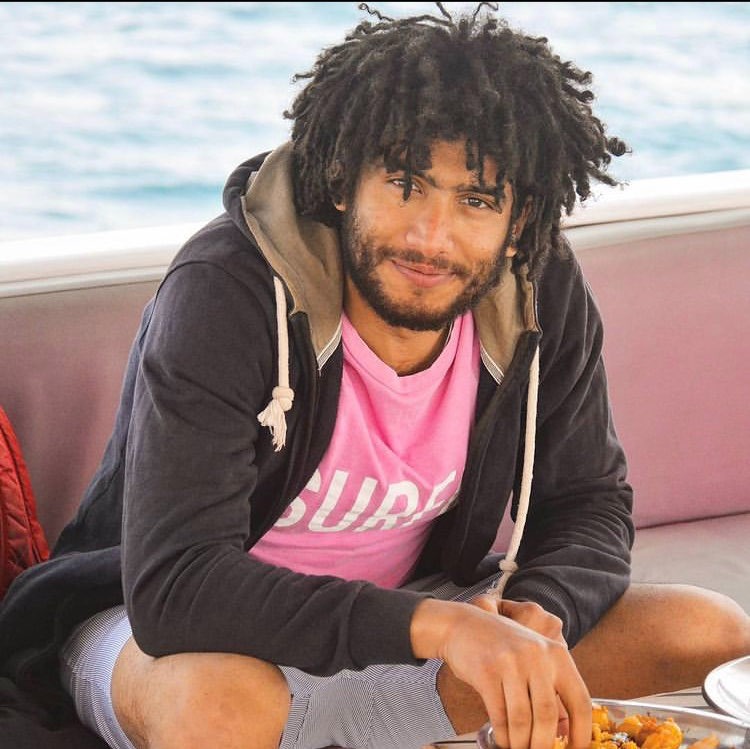 Badr Mohamed seated on a bench wearing a pink t-short and a black cardigan