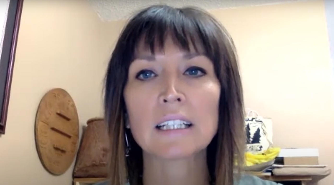 A woman with dark, shoulder-length brown hair and cropped fringe speaks into the camera