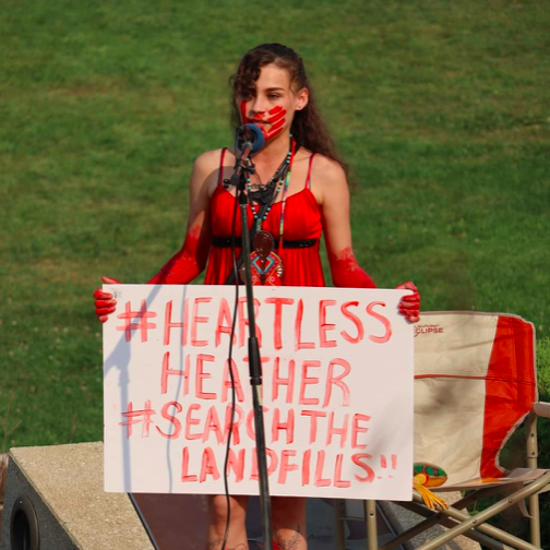 A young woman stands at a microphone wearing a red dress and a red handprint painted on her face while holding a sing that reads #HeartlessHeather #SearchTheLandfills
