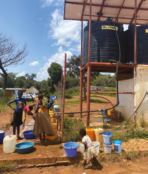 Two adults, one with a baby on her back, and two children use an outdoor well on a sunny day. 