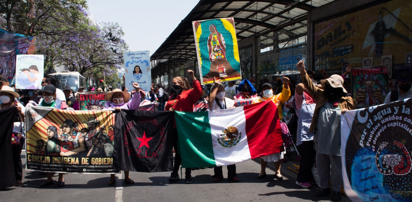 Protesters carrying flags and brightly coloured signs march on an asphalt road on a bright day.
