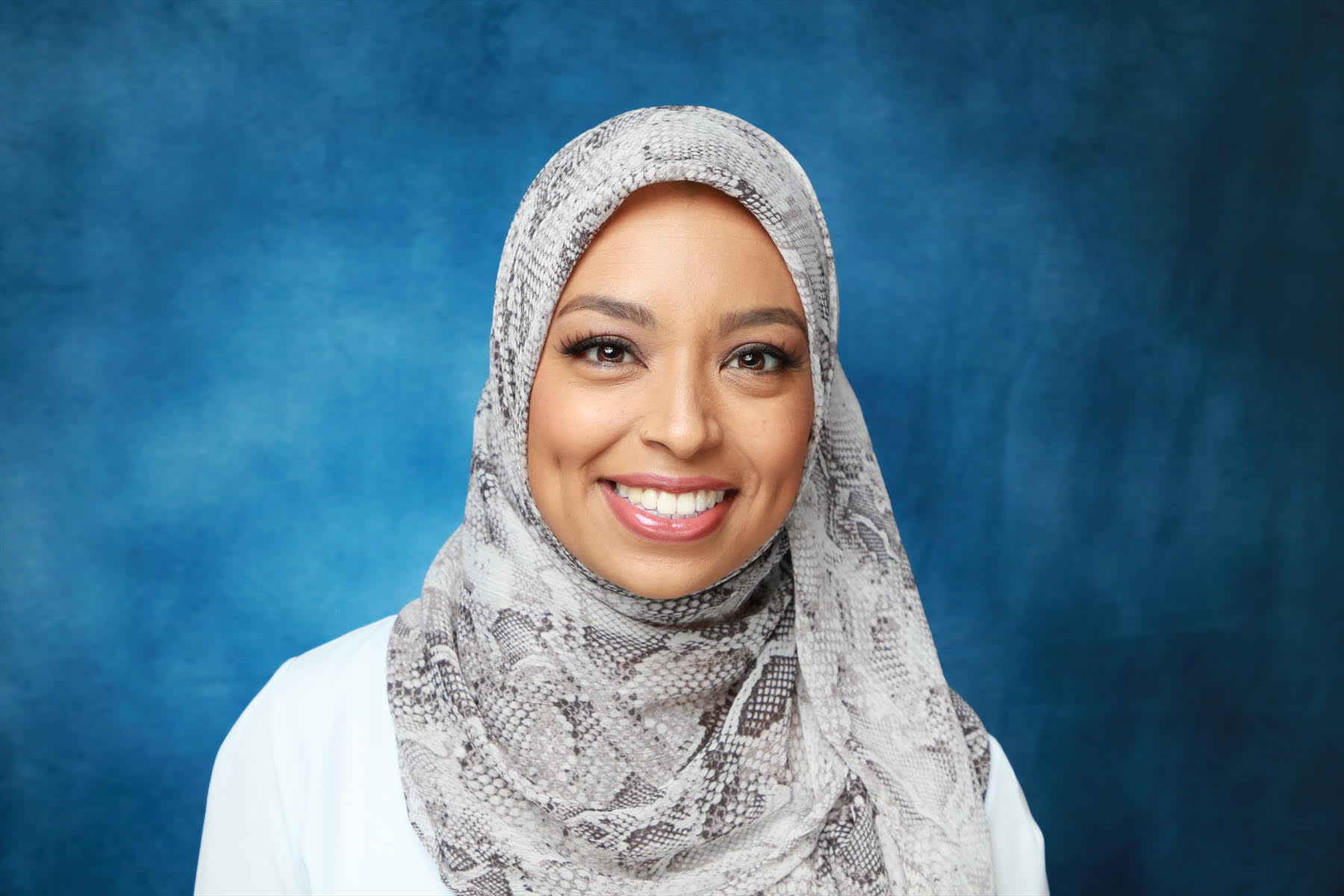 A smiling woman wearing a hijab with a grey and white snakeskin pattern.