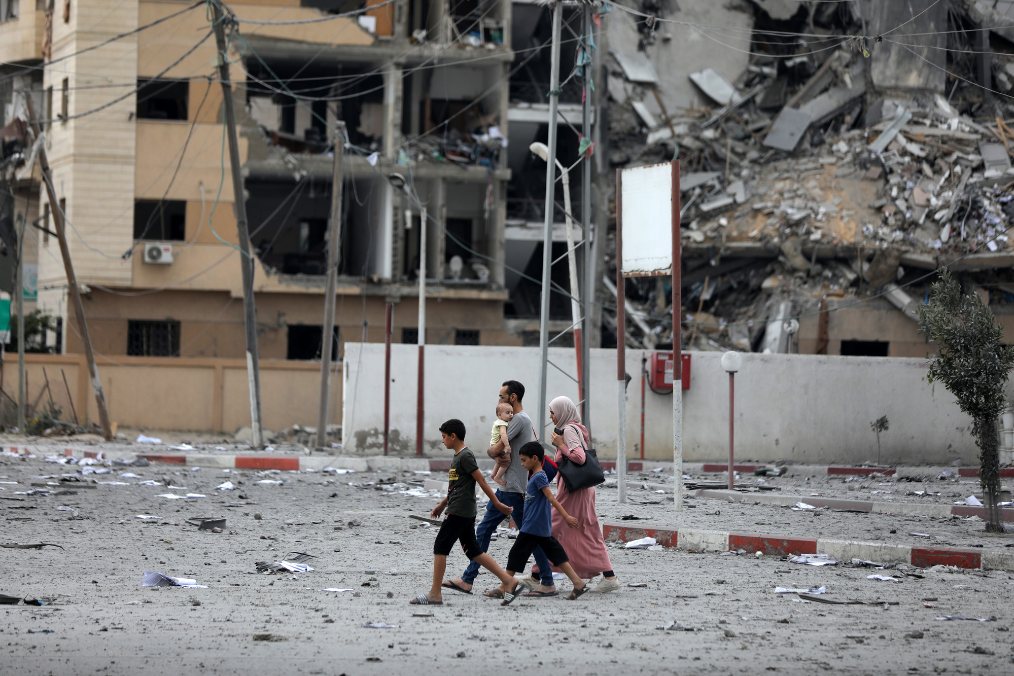 A woman, two boys, and a man carrying a baby walking on a road in front of a bombed-out apartment building.