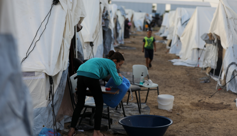A girl wearing a teal sweater and dark trousers pours water from a jug into a black plastic basin near a row of white tents at what appears to be a refugee camp.