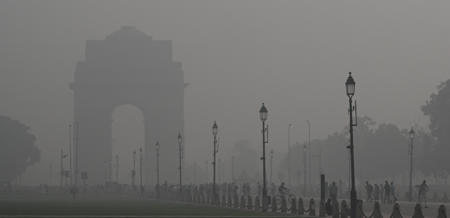 A thick layer of grey smog blankets an arch and street lamps on a street being used by cyclists and pedestrians
