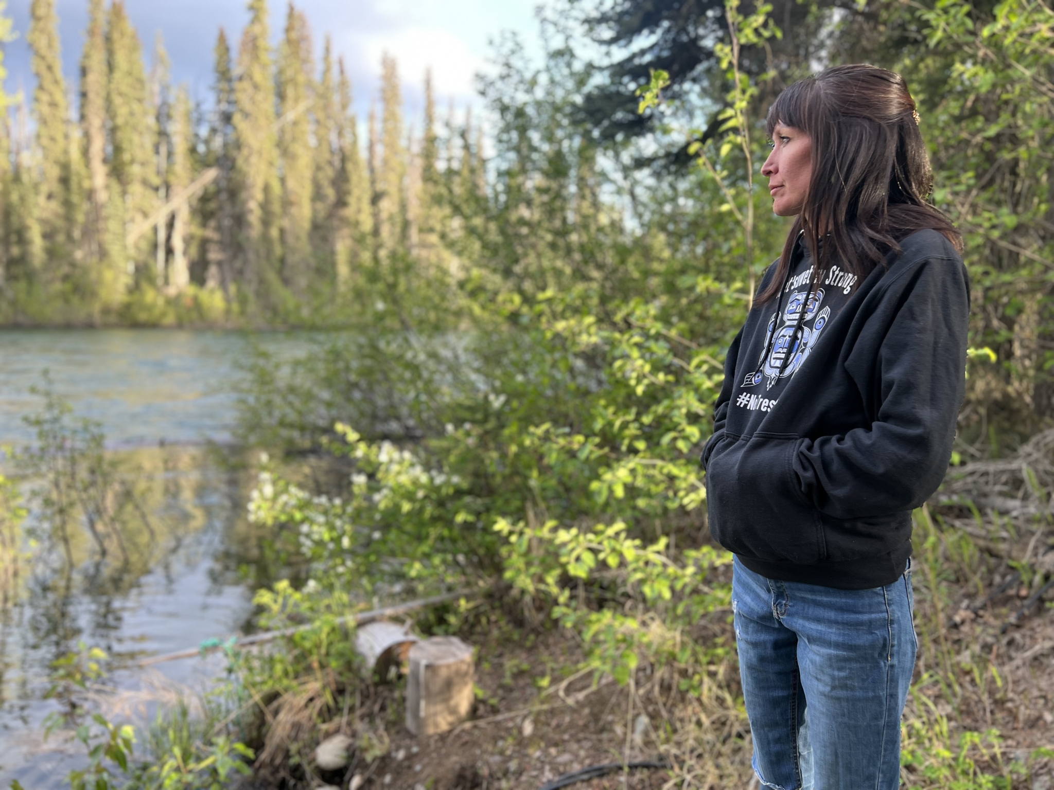 A woman wearing a black hoodie and blue jeans stands as she gazes toward a river. (Alli McCracken/Amnesty International)