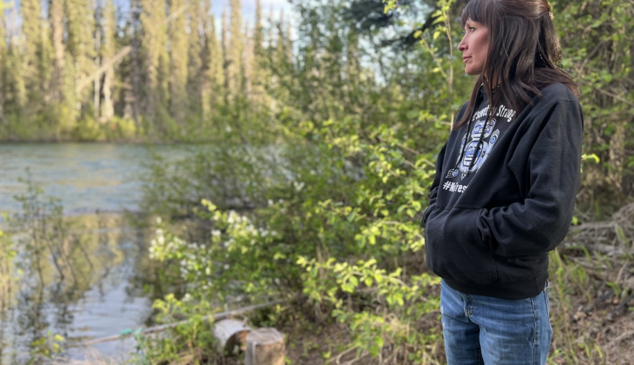 A woman wearing a black hoodie and blue jeans stands as she gazes toward a river. (Alli McCracken/Amnesty International)